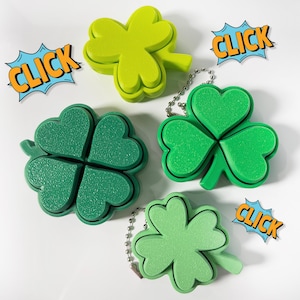 May include: Four green shamrock-shaped keychains in varying shades of green. Two have silver chains. Comic book style speech bubbles with the word "CLICK" in orange and yellow are also present. The background is white.