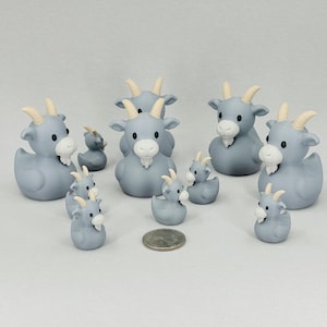 May include: A collection of gray goat-shaped rubber duckies with white beards and tan horns. The ducks vary in size, with some larger and some smaller. A US quarter is included for size comparison. The ducks are arranged on a white surface.