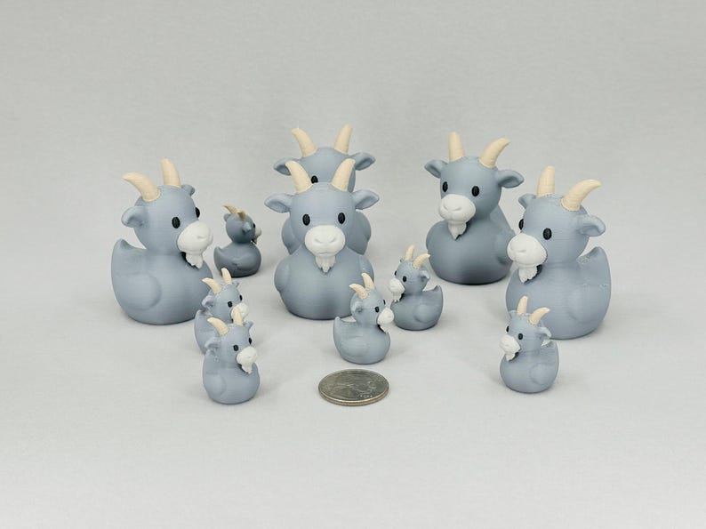 May include: A collection of gray goat-shaped rubber duckies with white beards and tan horns. The ducks vary in size, with some larger and some smaller. A US quarter is included for size comparison. The ducks are arranged on a white surface.