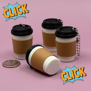 May include: Four miniature coffee cup keychains with black lids and brown sleeves, arranged on a pink surface. One cup is lying on its side. A coin is next to the cups. Comic book style speech bubbles with the word "CLICK" are in the image.