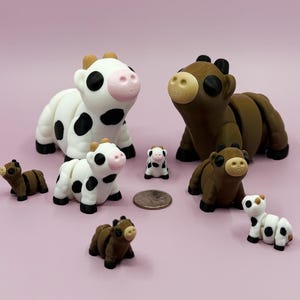 May include: A collection of handcrafted cow figurines in various sizes. The cows are white with black spots and brown with tan accents. A coin is included for size reference. The figurines are set against a pink background.