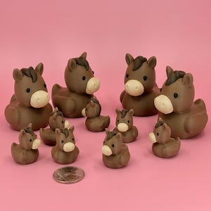 May include: A collection of brown horse figurines with white muzzles and dark manes. The figurines vary in size, with larger ones in the back and smaller ones in the front. A coin is included for scale, all set against a pink background.