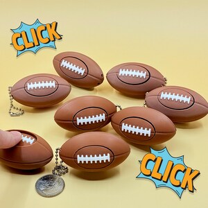 May include: Brown football-shaped keychains with white stitching, arranged on a yellow background. One keychain is being touched by a finger. The word "CLICK" is in a speech bubble.