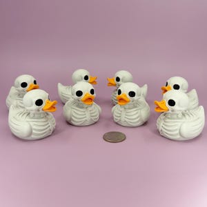 May include: A group of seven white rubber duck figurines with black eyes and orange beaks. The ducks have a skeleton design on their bodies. A coin is placed in the center of the ducks on a light purple surface.