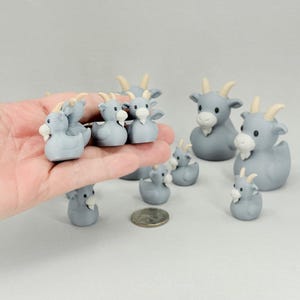 May include: A collection of gray goat figurines with white beards and yellow horns. The figurines vary in size and are arranged on a light gray surface. Some are held in a hand.