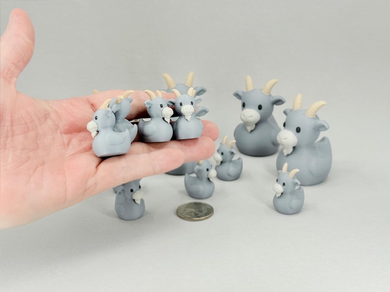 May include: A collection of gray goat figurines with white beards and yellow horns. The figurines vary in size and are arranged on a light gray surface. Some are held in a hand.