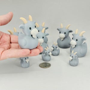 May include: A collection of gray goat figurines with cream-colored horns and beards. The figurines vary in size, with some held in a hand. A coin is next to the figurines. The goat figurines are a whimsical decorative item.