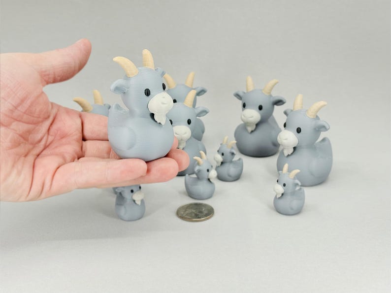 May include: A collection of gray goat figurines with cream-colored horns and beards. The figurines vary in size, with some held in a hand. A coin is next to the figurines. The goat figurines are a whimsical decorative item.