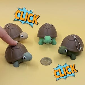 May include: Four turtle keychains with brown shells and green or tan heads and feet are arranged on a yellow surface. A finger points at one turtle. The word "CLICK" is in a blue and orange comic book style speech bubble.