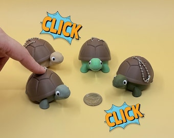 May include: Four turtle keychains with brown shells and green or tan heads and feet are arranged on a yellow surface. A finger points at one turtle. The word "CLICK" is in a blue and orange comic book style speech bubble.