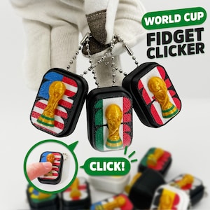 May include: Three rectangular black fidget clickers with metal ball chains, each featuring a gold-colored World Cup trophy emblem over different national flag designs. The text WORLD CUP FIDGET CLICKER and CLICK! is visible in the image.