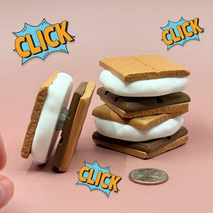 May include: A stack of s'mores with graham cracker, marshmallow, and chocolate layers. The image includes the word "CLICK" in a comic book style speech bubble. A quarter is next to the s'mores.
