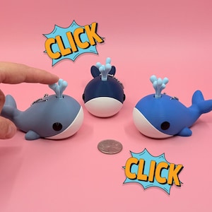 May include: Three whale-shaped keychains in shades of blue, each with a white belly and a water spout design. The word "CLICK" is in a speech bubble. A hand is touching one of the whales.