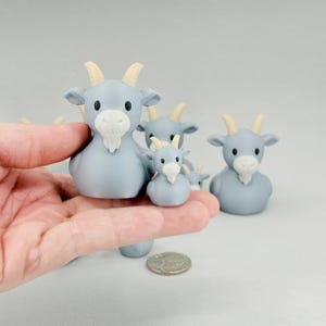 May include: A collection of light blue goat figurines with cream-colored horns and beards. The figurines vary in size, with one held in a hand. The goats have black eyes and are arranged on a light gray surface.