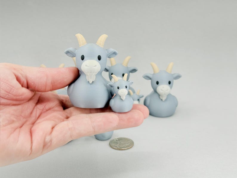 May include: A collection of light blue goat figurines with cream-colored horns and beards. The figurines vary in size, with one held in a hand. The goats have black eyes and are arranged on a light gray surface.