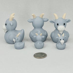 May include: A collection of light blue goat figurines with cream-colored horns and beards. The set includes three larger goats and three smaller ones, with a quarter coin for size comparison. The goats have a whimsical, cartoonish design.
