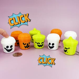 May include: A collection of Halloween-themed miniature cups in orange, white, and green, each with a different jack-o'-lantern or ghost face. The word "CLICK" is in a comic book style speech bubble. A US quarter is next to the cups.