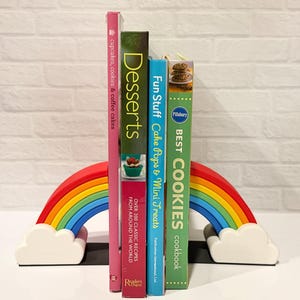 May include: A colorful rainbow bookend supports a collection of cookbooks. The books include titles like "Cupcakes, Cookies & Coffee Cakes," "Desserts," "Fun Stuff Cake Pops & Mini Treats," and "Best Cookies." The rainbow bookend adds a playful touch to the display.