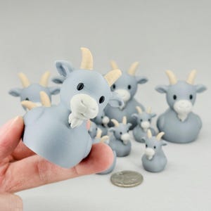 May include: A collection of light blue goat figurines with cream-colored horns and white beards. The figurines vary in size, with some held in a hand. A coin is also present.
