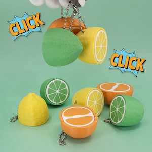 May include: A collection of fruit-shaped keychains, including lemons, limes, and oranges, in various forms: whole, halved, and sliced. The keychains are yellow, green, and orange, with silver chain attachments. The word "CLICK" is displayed in a comic book style.
