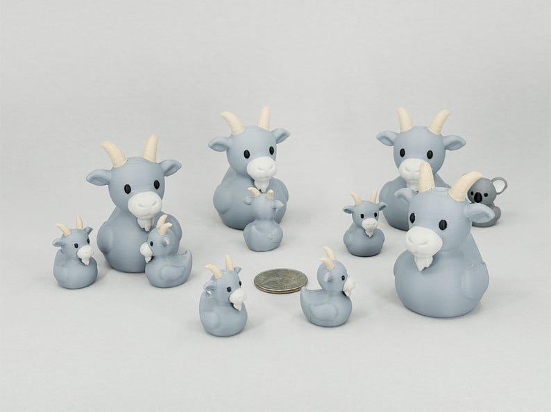 May include: A collection of light blue goat figurines with white beards and horns. The figurines vary in size, with some appearing to be baby goats. A small koala bear figurine is also present. A US quarter is included for size reference.