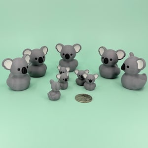 May include: A collection of gray koala figurines with white-lined ears and black eyes. The koalas vary in size, with several small ones grouped together. A coin is included for size reference. The background is a solid light green.