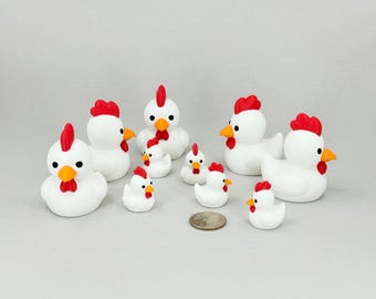 May include: A collection of white chicken figurines with red combs and yellow beaks. The figurines vary in size, with some standing upright and others in a sitting position. A coin is included for size reference.