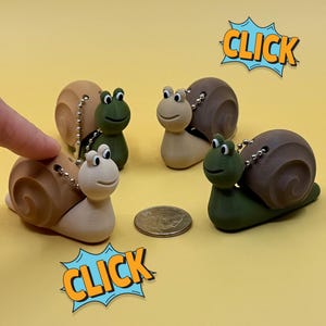 May include: A collection of snail-shaped keychains in various colors, including tan, brown, and green. Each keychain has a silver chain and a smiling face. The word "CLICK" is in a blue and yellow comic-style bubble.