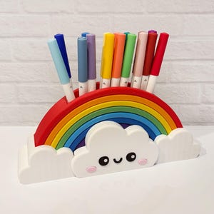 May include: A rainbow-shaped desk organizer featuring a white cloud base with a smiling face and pink cheeks. The organizer has multiple slots holding various colored felt-tip pens in shades of blue, purple, yellow, orange, green, pink, and red.