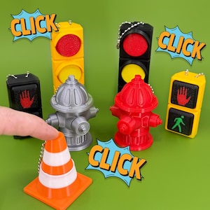 May include: A set of five colorful, miniature traffic light and fire hydrant keychains. The keychains are made of plastic and have a click sound when pressed. The keychains are arranged on a green surface with a traffic cone keychain in the foreground.