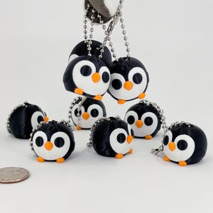 May include: A collection of small, round penguin charms with black bodies, white faces, and orange beaks and feet. The penguin charms are attached to silver ball chains. A U.S. quarter is in the image for size comparison.