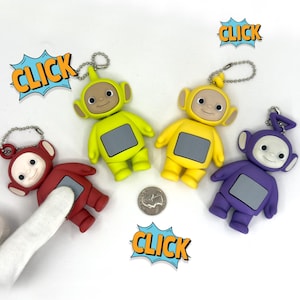 May include: Four Teletubbies keychains in red, yellow, green, and purple. Each figure has a screen on its chest and a silver keychain. Comic book style "CLICK" text bubbles are in the background.