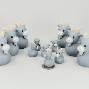 May include: A collection of gray goat-shaped figurines with white beards and horns. The figurines vary in size, with some resembling duck shapes. A coin is placed near the smallest figurine for size comparison.