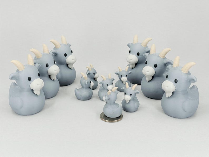 May include: A collection of gray goat-shaped figurines with white beards and horns. The figurines vary in size, with some resembling duck shapes. A coin is placed near the smallest figurine for size comparison.