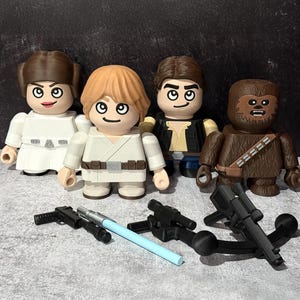 May include: Four character figures from the Star Wars franchise, including Princess Leia, Luke Skywalker, Han Solo, and Chewbacca. Also included are several black blasters and a light blue lightsaber. The figures are standing on a gray surface.
