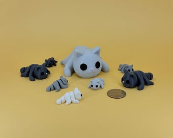 May include: A collection of small cat figurines in various colors, including gray, dark gray, and white, are arranged on a yellow surface. The figurines vary in size, with a larger gray cat in the center. A coin is also present.