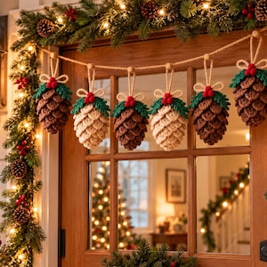 May include: A garland of six crocheted pinecone ornaments hanging from a jute string. Each pinecone features brown or cream yarn, green crocheted leaves, and three small red pom-poms at the top, suspended by a loop of matching jute twine.