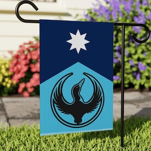 May include: A garden flag featuring a design with a black bird emblem inside a circle, a white star, and a blue and light blue color scheme. The flag is hanging on a black metal stand in a garden setting.