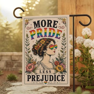 May include: A garden flag with the text "MORE PRIDE LESS PREJUDICE" and a portrait of a person wearing rainbow heart-shaped sunglasses. The flag is decorated with flowers and leaves, and is displayed on a black metal stand.