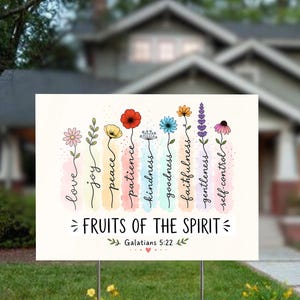 May include: A white rectangular sign with the words "Fruits of the Spirit" and "Galatians 5:22". The sign features colorful flowers with words like "love", "joy", and "peace". The sign is displayed on a lawn with a house in the background.