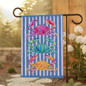 May include: A blue and white striped garden flag featuring three colorful crabs. The crabs are painted in shades of pink, green, and yellow, with detailed claws and bodies. The flag is displayed on a black metal stand in a garden setting.