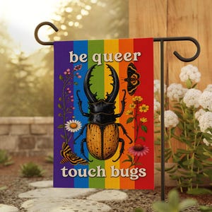 May include: A garden flag with a rainbow background and the words "be queer touch bugs". The flag features a large beetle illustration with butterflies and flowers. The flag is displayed on a black metal stand.