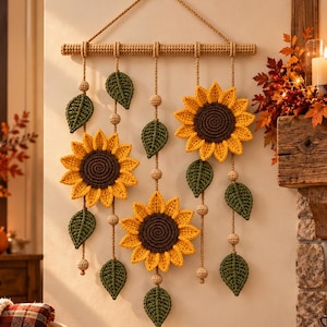 Könnte beinhalten: Ein Wandbehang mit drei gehäkelten Sonnenblumen mit dunkelbraunen Mitten und leuchtend gelben Blütenblättern, durchsetzt mit grünen gehäkelten Blättern und kleinen Holzperlen, aufgehängt an einem horizontalen Holzstab mit einer Kordel.