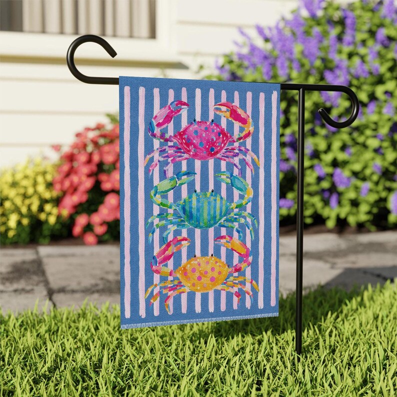May include: A decorative garden flag featuring a blue and white striped background with four colorful crab illustrations. The crabs are in shades of pink, green, blue, and yellow. The flag is displayed on a black metal stand.
