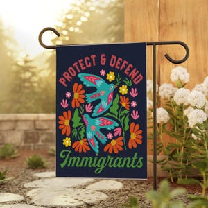 May include: A navy blue garden flag with the words "PROTECT & DEFEND Immigrants" in red and green. The flag features two turquoise birds with red accents, surrounded by orange, pink, and yellow flowers and green foliage.