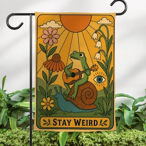 May include: A garden flag featuring a whimsical illustration. A green frog plays a guitar while riding a snail, with a sun, flowers, and the text "STAY WEIRD". The flag is displayed on a black metal stand.