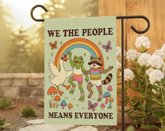 Puede incluir: Una bandera de jardín con el texto "WE THE PEOPLE MEANS EVERYONE" y una ilustración retro de un ganso, una rana y un mapache bailando bajo un arcoíris. La bandera es de color verde claro con gráficos coloridos y se muestra en un soporte de metal negro.