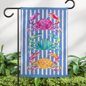 May include: A blue and white striped garden flag featuring three colorful watercolor crabs. The crabs are pink, blue, and yellow, with detailed claws and bodies. The flag is hanging on a black metal stand, perfect for outdoor decor.