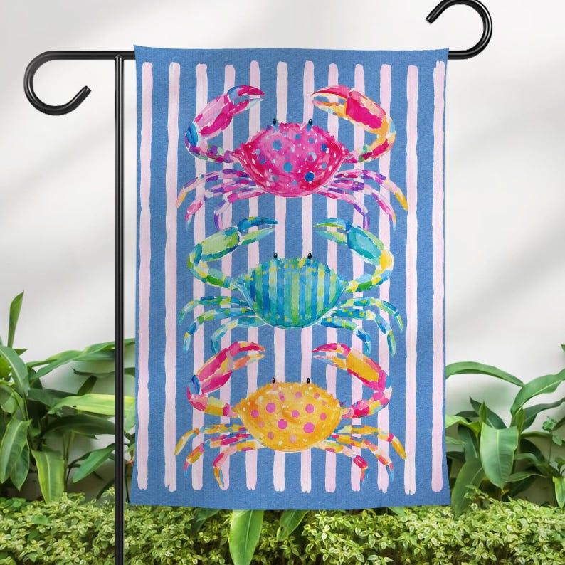 May include: A blue and white striped garden flag featuring three colorful watercolor crabs. The crabs are pink, blue, and yellow, with detailed claws and bodies. The flag is hanging on a black metal stand, perfect for outdoor decor.