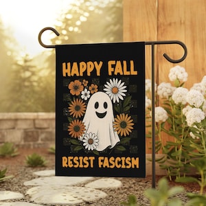 May include: Black garden flag with the text "HAPPY FALL" and "RESIST FASCISM" in orange. The flag features a smiling ghost surrounded by orange and white flowers against a checkered background.